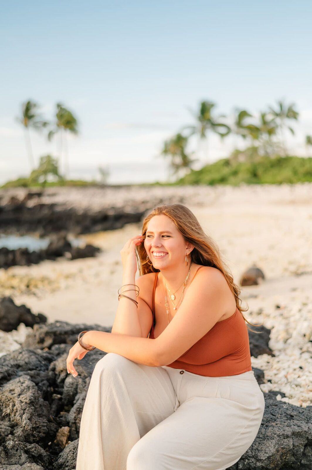 A woman with long hair sits on a rock at a beach, smiling and looking into the distance. She wears a brown sleeveless top and light pants. The scene resembles Big Island senior portraits, with palm trees, rocks, and a clear sky painting the perfect backdrop.