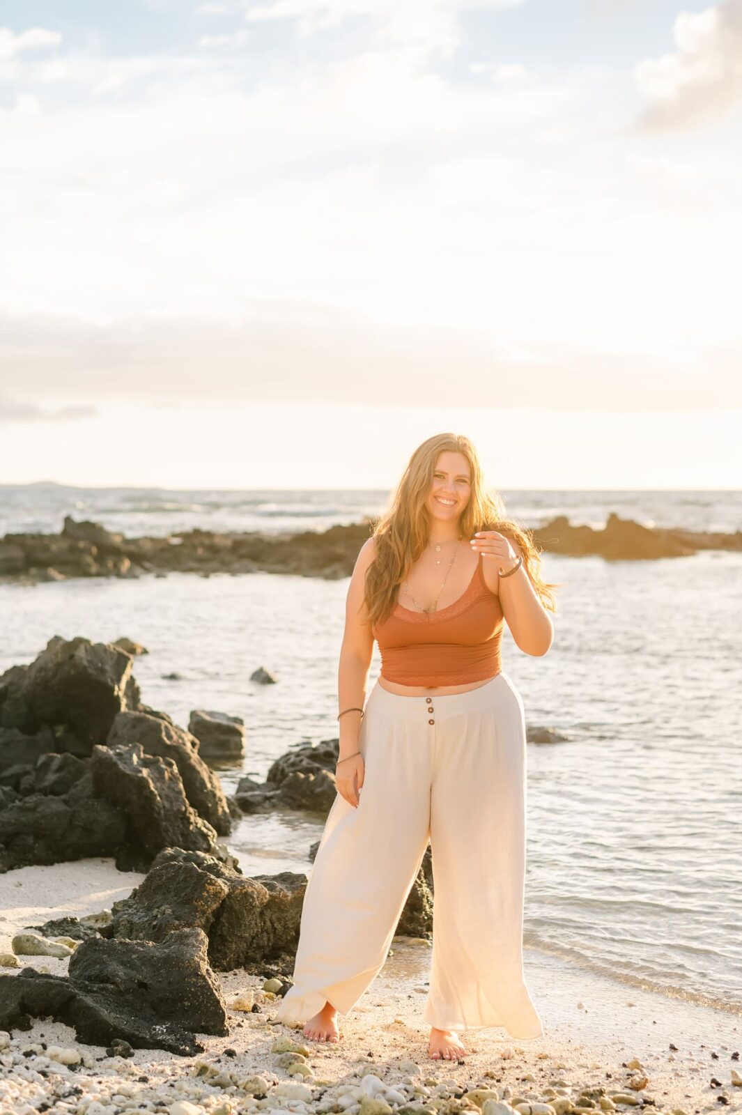 On a rocky beach, a woman smiles at the camera in her rust-colored top and white pants. Her long hair flows as the sun shines brightly over the sea and sky. Its a perfect setting for Big Island senior portraits, capturing natures beauty in every frame.