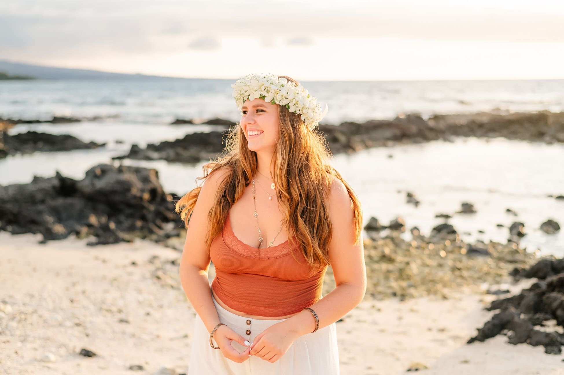 A woman stands on a rocky beach, adorned in a flower crown and orange top, captured in an enchanting Big Island senior portrait. She smiles softly, gazing into the bright sky with the vast ocean stretching out behind her.