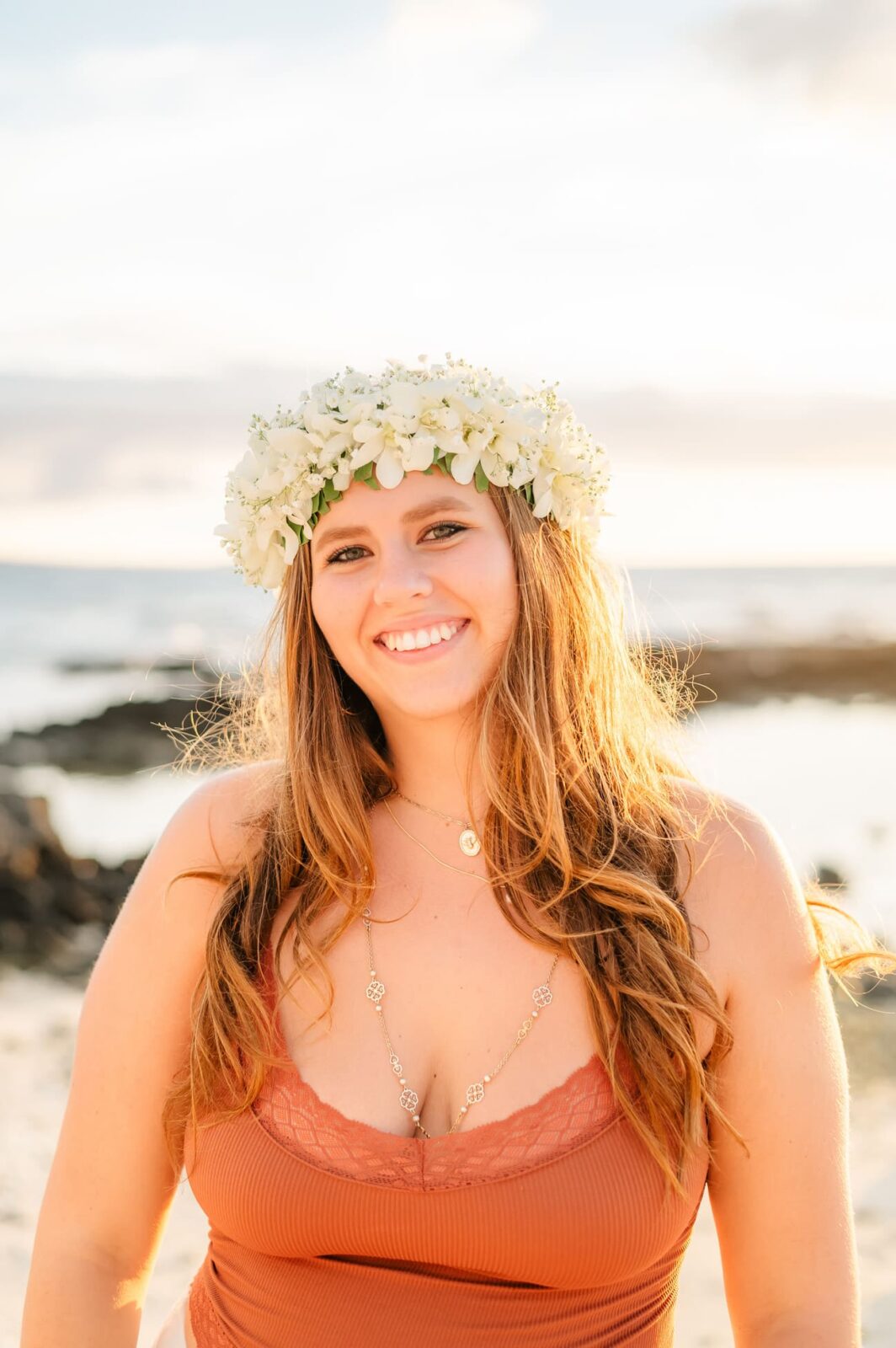 Smiling woman wearing a floral crown and a coral top with a lace trim, captured in her Big Island senior portrait. She stands on the beach at sunset, with a rocky shoreline and calm sea under a soft, cloudy sky.