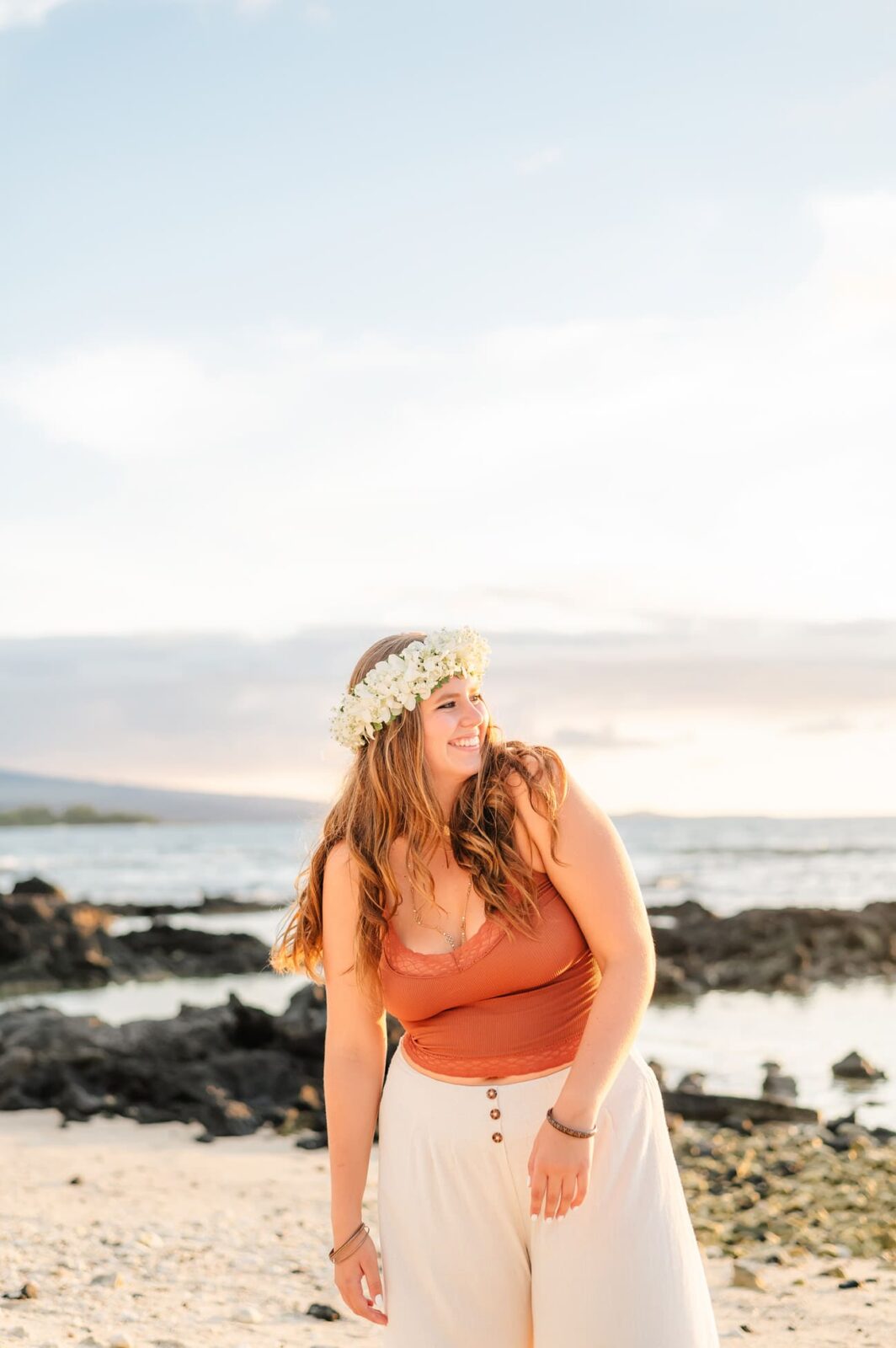 A person stands on a Big Island beach wearing a white flower crown, an orange top, and cream pants. They are smiling and looking to the side, with a backdrop of rocks, ocean, and a partly cloudy sky during sunset—an ideal setting for senior portraits.