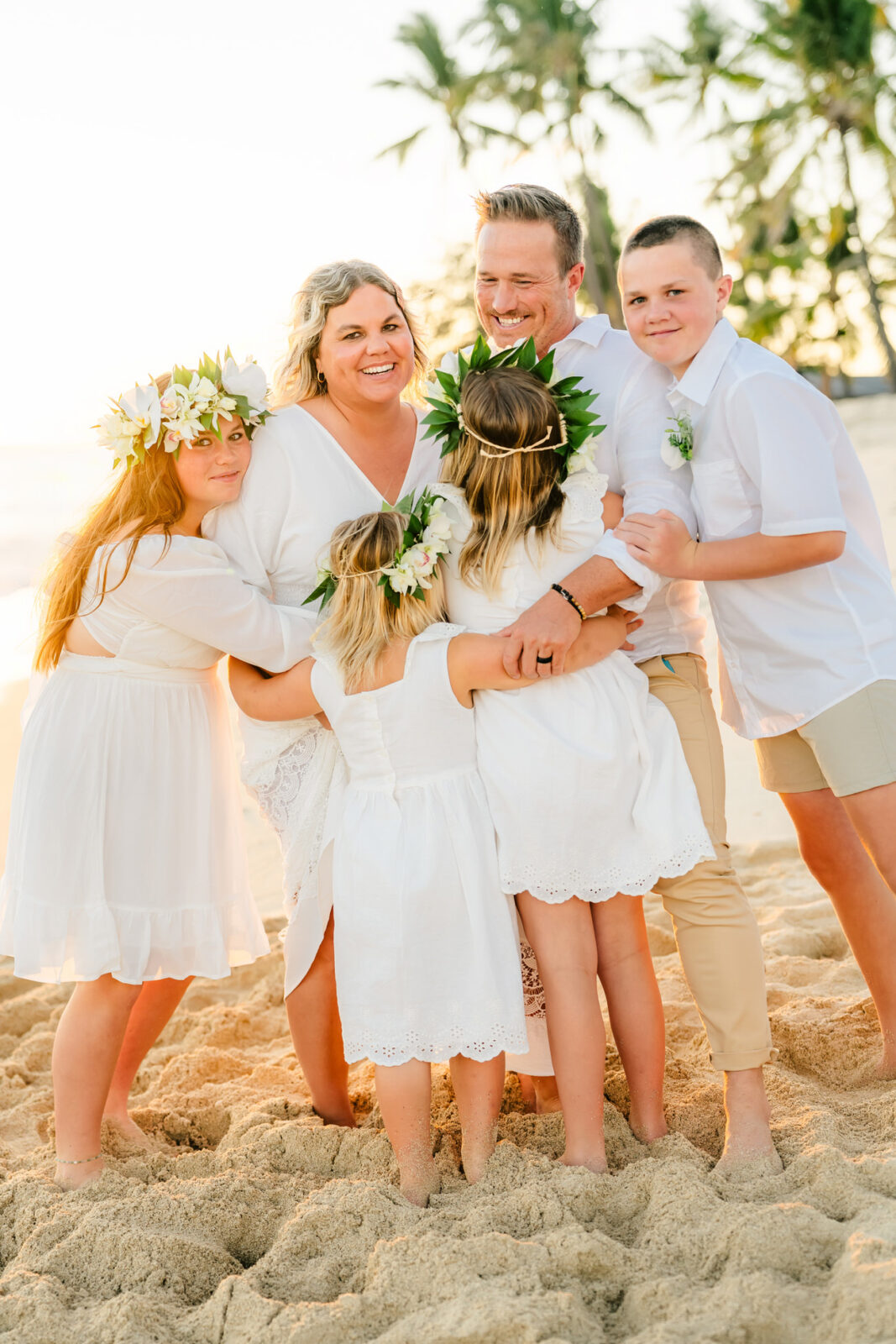 A family of six, dressed in white, stands happily on a sandy beach at sunset in Kapolei. They embrace with palm trees and the ocean as their backdrop. The children wear floral crowns, capturing the joyful and serene mood of this unforgettable family photo.