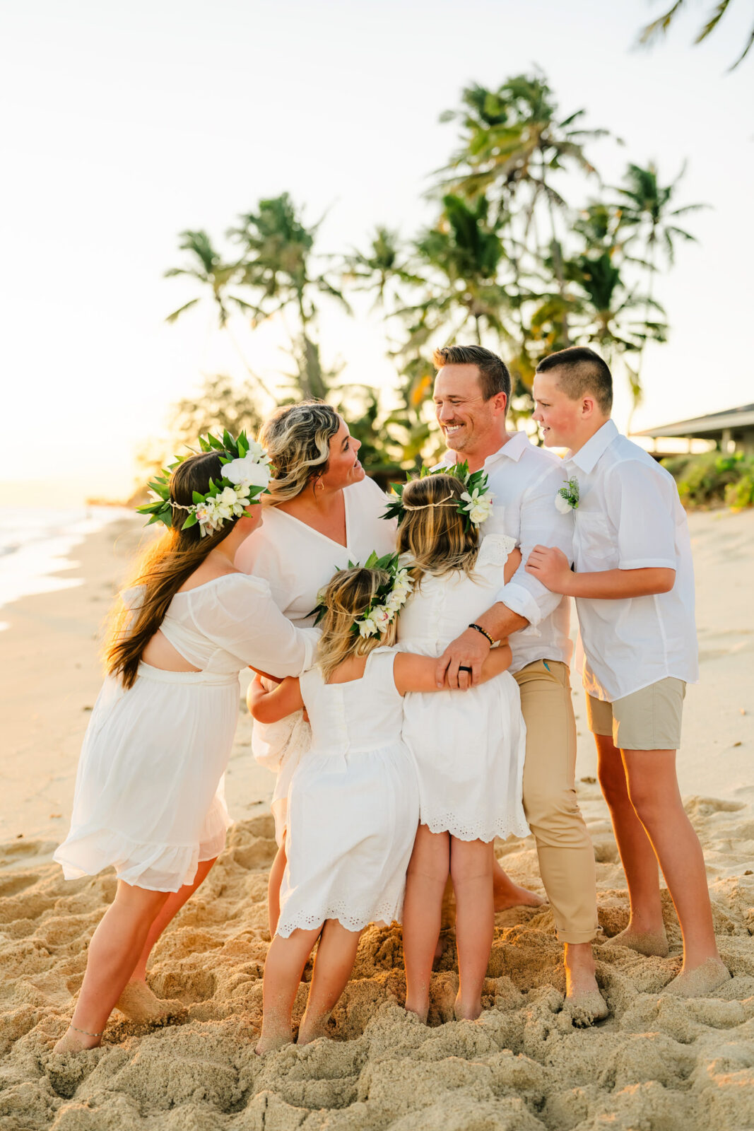 A family of five, all dressed in white, joyfully embrace on a sandy beach in Kapolei. The sun sets behind them, illuminating palm trees and the ocean. This idyllic moment captures the warmth and happiness typical of family photos taken in such a beautiful setting.