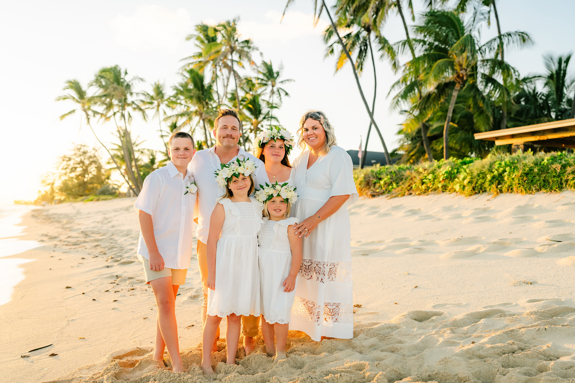 A family of five enjoys a sandy beach at sunset in Kapolei, dressed in white outfits. The two girls wear flower crowns as palm trees and lush greenery create a stunning backdrop for their cherished family photos.