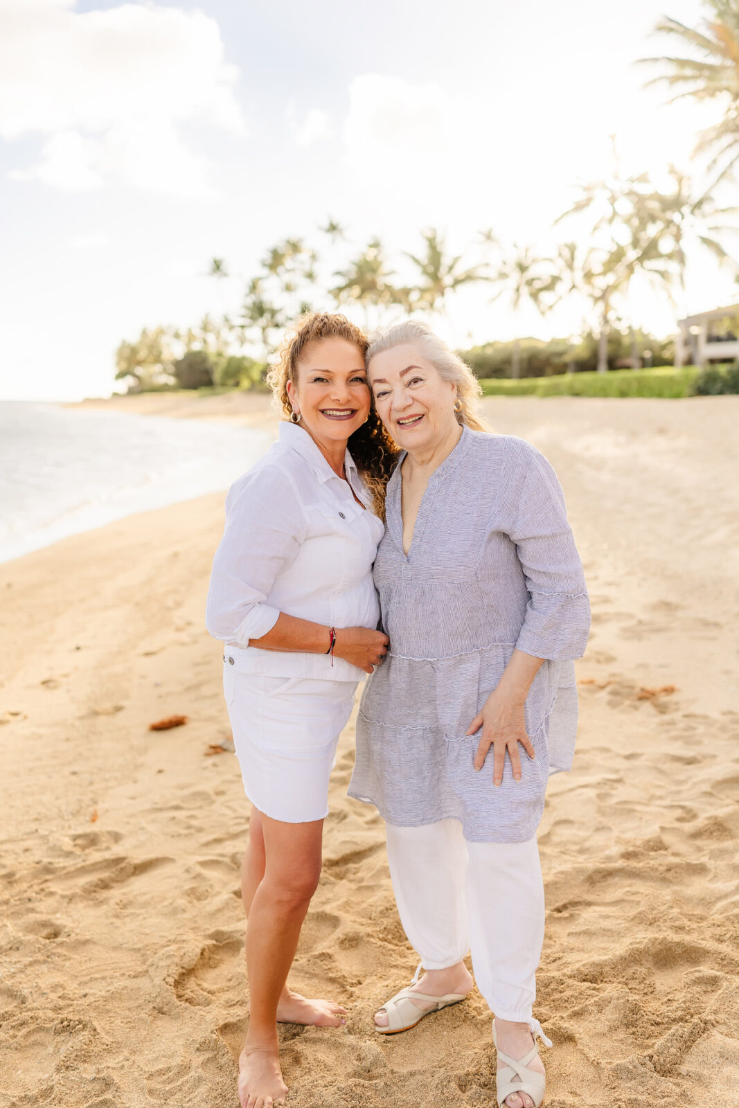 Two women stand on a sandy beach, smiling at the camera. One is wearing a white outfit, and the other is in a light blue top and white pants. Palm trees and ocean waves are visible in the background under a sunny sky.