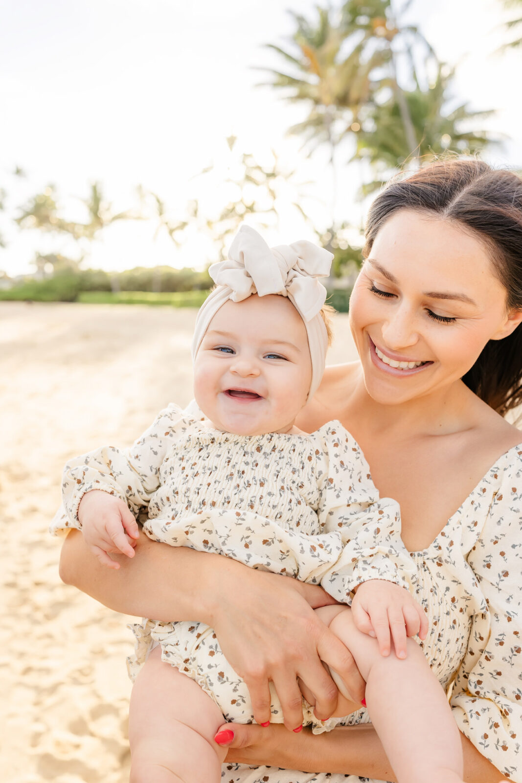 A woman holding a smiling baby at the beach. Both are wearing matching floral outfits. The woman gazes affectionately at the baby, who has a large bow headband. The background features sand, palm trees, and a clear sky.