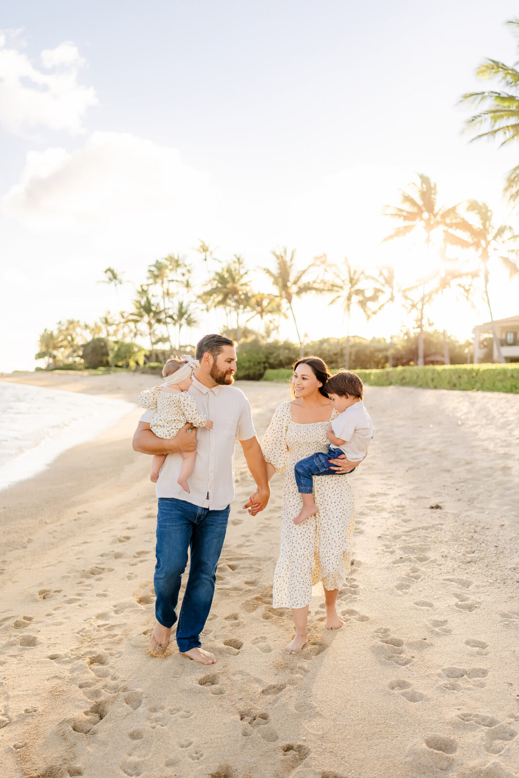 A family of four walks along a sandy beach at sunset. The parents are holding hands, each carrying a young child. The sky is bright with scattered clouds, and palm trees are visible in the background.