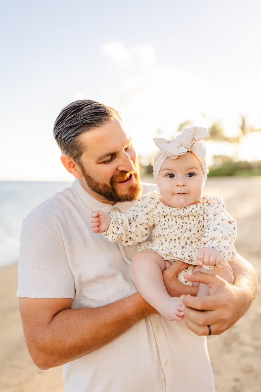 A bearded man holding a baby on a sunny beach. The baby is wearing a headband and a patterned outfit, looking at the camera. The man, smiling, is wearing a light shirt. The background shows the sea and palm trees.