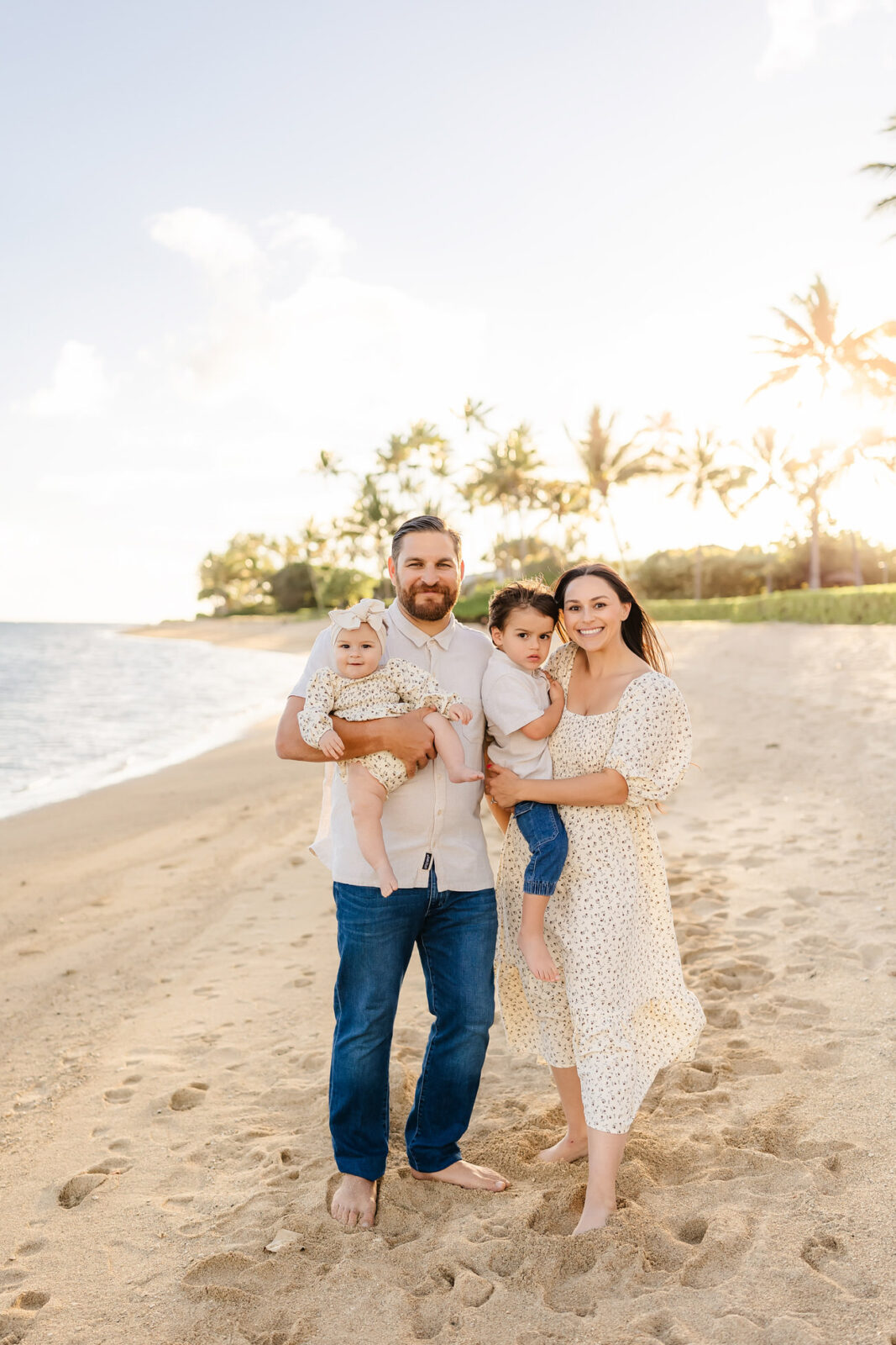 A family of four stands on a sandy beach at sunset. The man holds a baby, and the woman holds a toddler. They are all dressed in light-colored clothing, with palm trees and the ocean in the background. The sun is setting behind them.