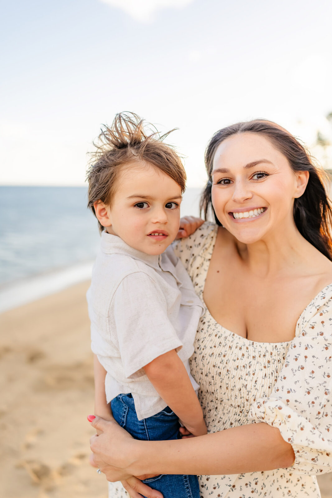 Smiling woman holding a young boy on a sandy beach. The woman has long, dark hair and is wearing a floral dress. The boy has tousled hair, wears a light shirt and jeans, and gazes at the camera. The ocean and sky are in the background.