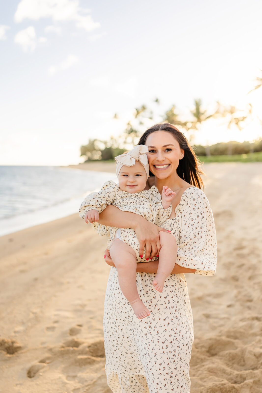 A woman holding a baby stands on a sandy beach. Both are smiling and wearing matching light-colored outfits with small floral patterns. The sun is setting in the background, and the ocean and palm trees are visible along the shoreline.