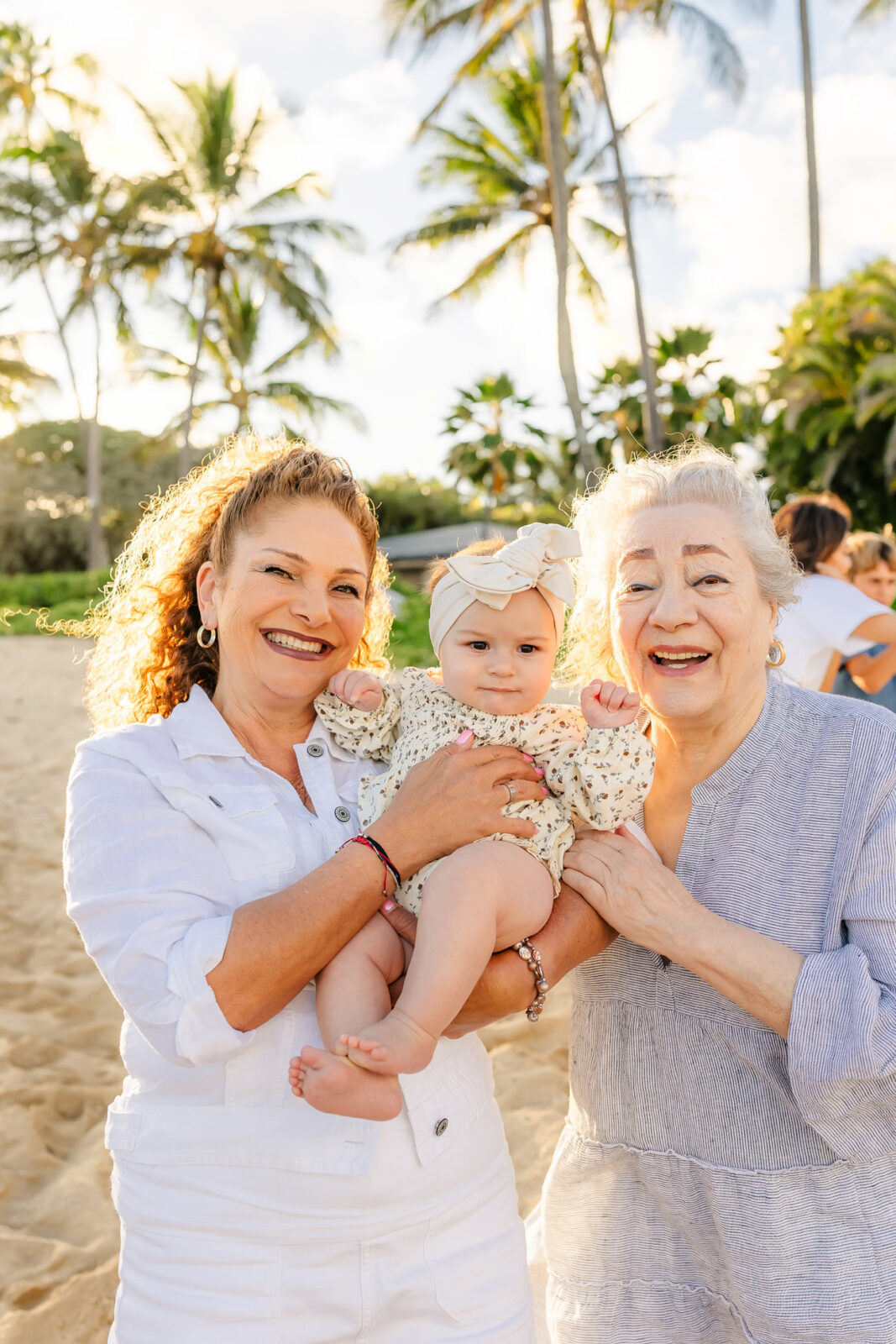 Two women, one in white and one in gray, smile while holding a baby wearing a floral outfit and bow on a beach. Palm trees and a clear sky are in the background, with people visible in the distance.