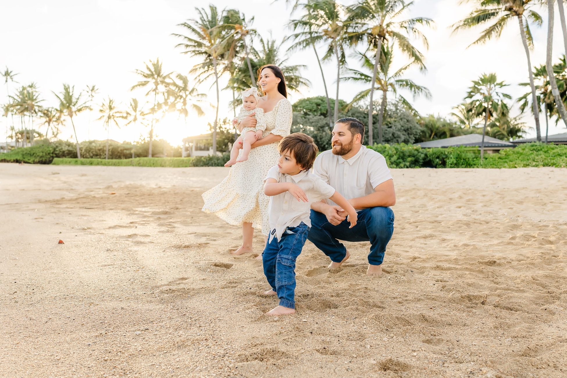 A family at a beach during sunset. A woman holding a baby stands next to a man crouching on the sand, while a young boy plays nearby. They are surrounded by palm trees and the sky is partly cloudy.