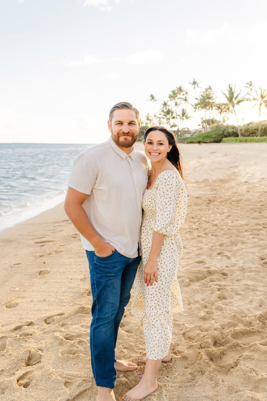 A couple stands on a sandy beach with the ocean and palm trees in the background. The man is wearing a light-colored shirt and jeans, while the woman is in a patterned dress. They are smiling and facing the camera.