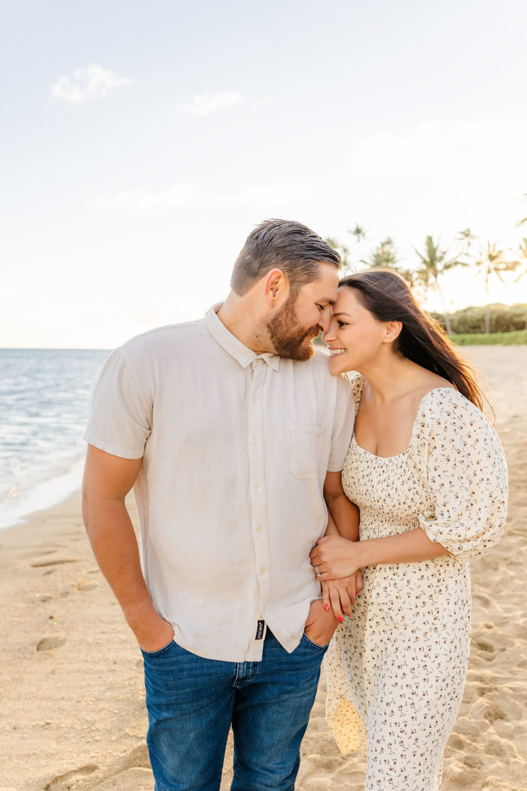 A couple stands on a sandy beach, smiling and leaning their foreheads together. The man wears a beige shirt and jeans, while the woman is in a white floral dress. Palm trees and the ocean are in the background under a clear sky.