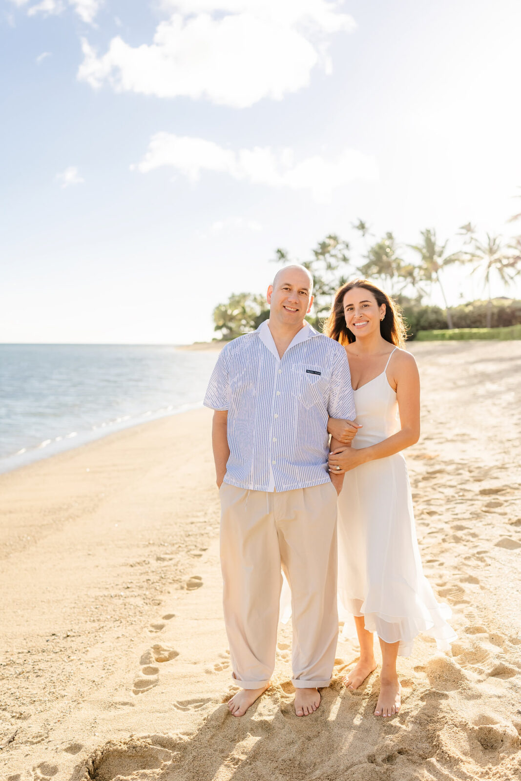 A couple stands on a sandy beach. The man wears a light blue striped shirt and beige pants, while the woman is in a white dress. They are barefoot, with calm ocean waves and palm trees in the background under a sunny sky.