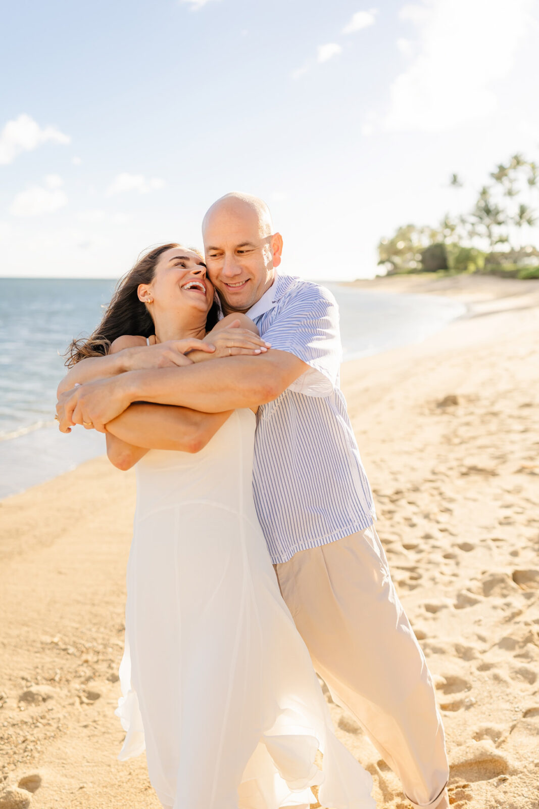 A couple standing on a sunny beach, smiling and embracing. The woman wears a white dress, and the man wears a striped shirt and beige pants. The sea and sky are visible in the background with scattered clouds and distant palm trees.