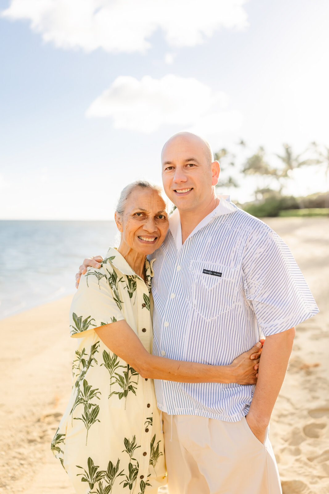 An elderly woman in a yellow floral dress and a man in a blue striped shirt smile and embrace on a sunny beach. The ocean and palm trees are visible in the background under a clear sky.