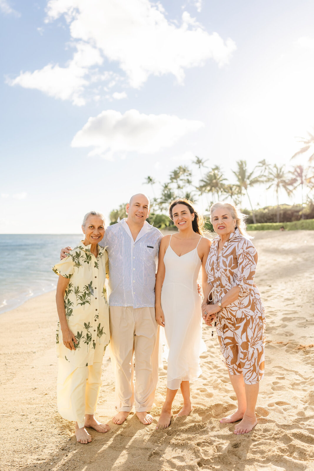 Four people standing closely on a sunny beach with gentle waves and palm trees in the background. They are barefoot, smiling, and wearing light, summery clothes. The sky is blue with a few clouds.