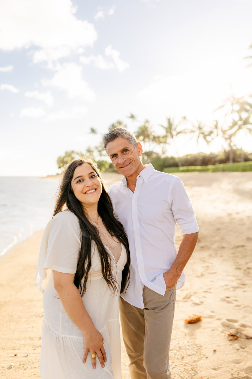 A man and woman stand smiling on a sandy beach under a bright sky. They are dressed in white, with palm trees and the ocean in the background.