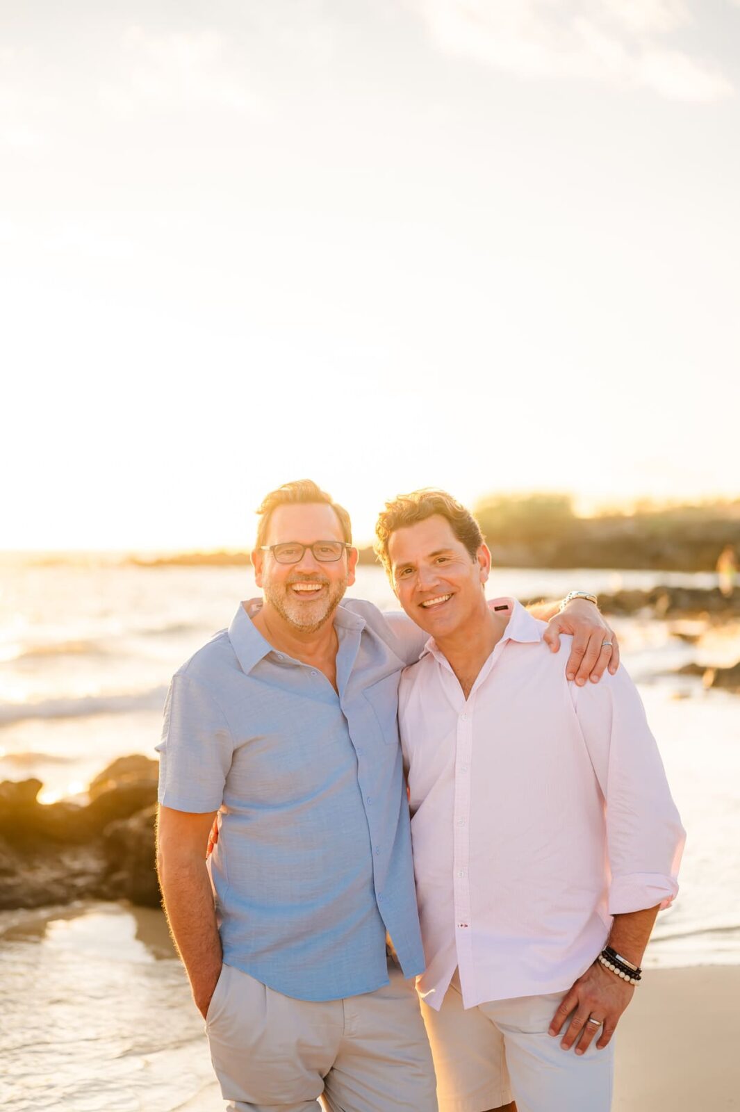 Two men stand close together on a beach at sunset, smiling at the camera during their Hawaii photo session with kids. One has his arm around the others shoulders. Both are dressed in light-colored clothing, with the ocean and rocks in the background.