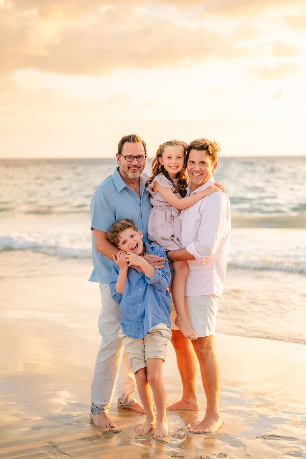 Two men and two children, a boy and a girl, stand barefoot on a beach at sunset, smiling during a Hawaii photo session with kids. One man holds the girl while the boy hugs the other man, with the ocean and golden sky behind them.