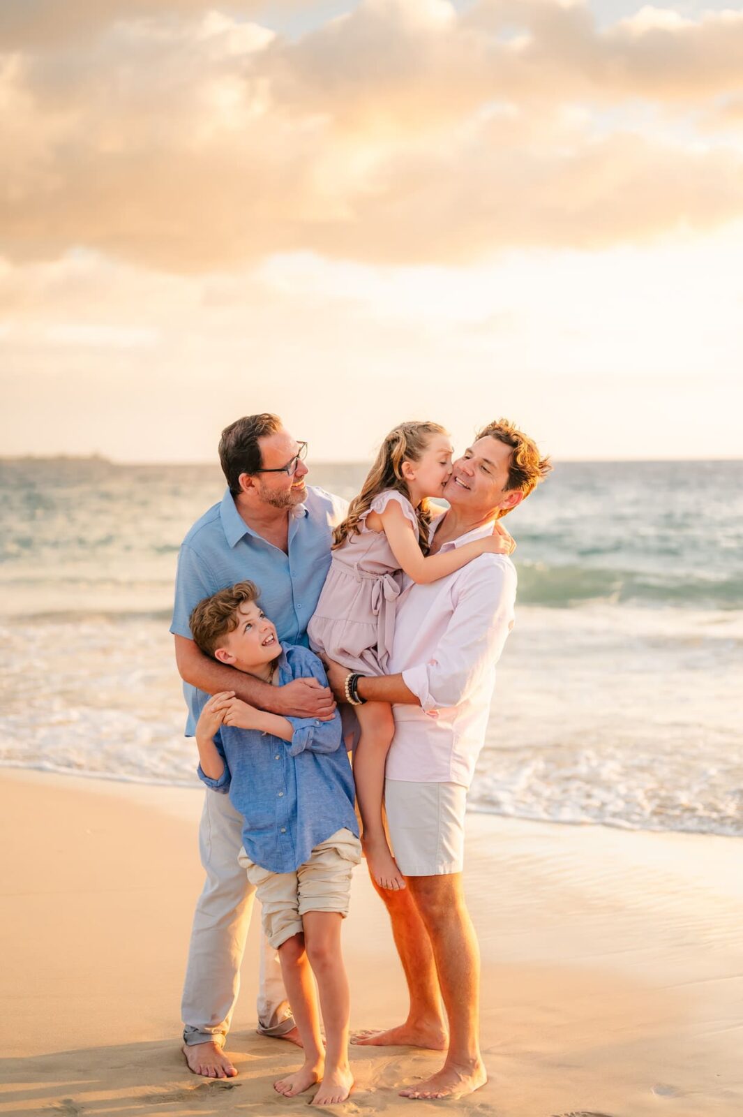 Two men stand barefoot on the beach at sunset during a Hawaii photo session with kids, smiling as the boy hugs one man’s leg and the girl laughs in the other man’s arms near the ocean waves.