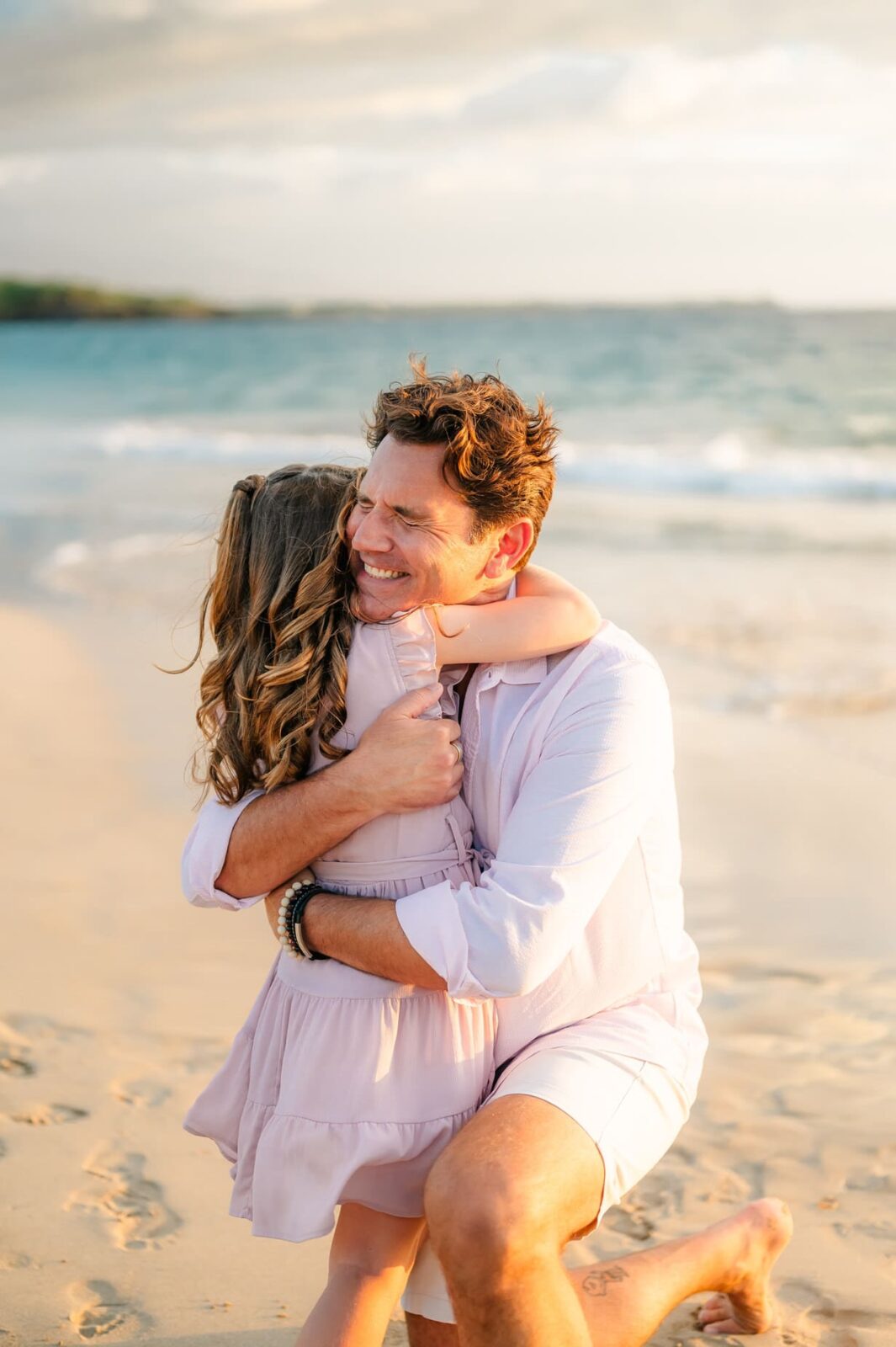 A man kneels on a sandy beach during a Hawaii photo session with kids, smiling as he hugs a young girl with long hair. Both wear light-colored clothes, and gentle ocean waves roll in the background.