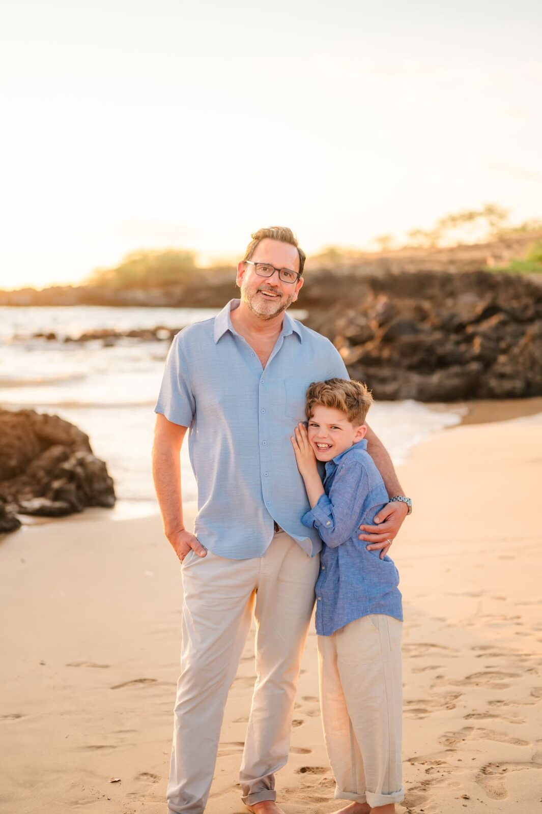 A smiling man and a young boy stand together on a sandy beach near rocks at sunset, both in light blue shirts and light pants. The boy leans affectionately against the man during their Hawaii photo session with kids.