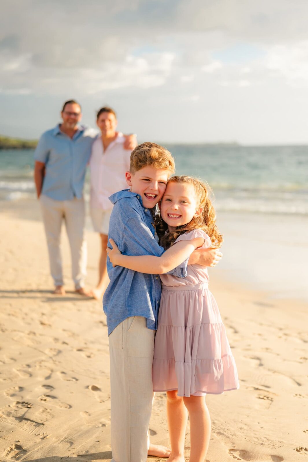 Two smiling children, a boy and a girl, hug each other on a sunny beach during a Hawaii photo session with kids, while two adults stand in the background near the shoreline with the ocean and sky behind them.