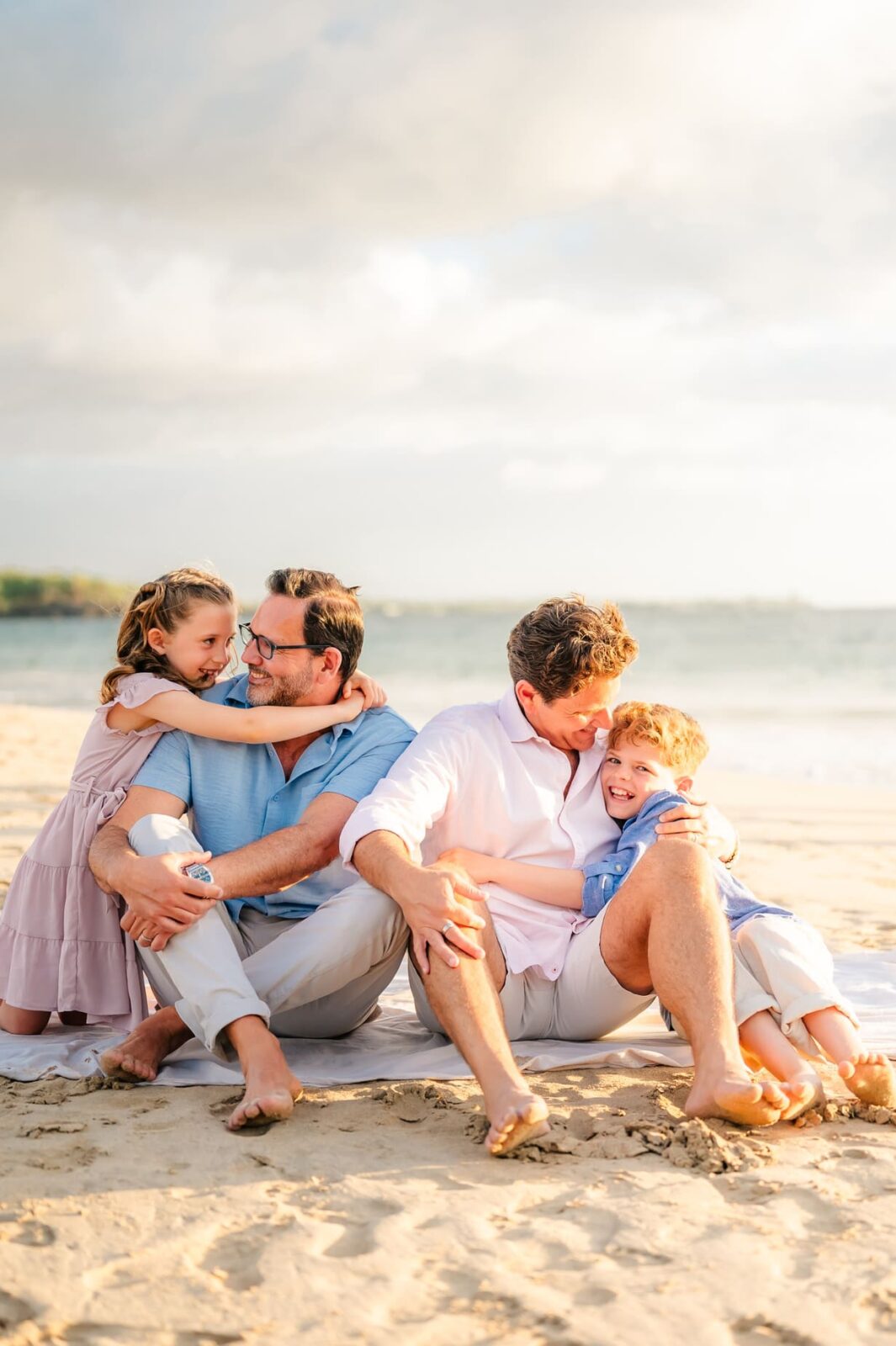Two men sit on a sandy beach during a Hawaii photo session with kids, a young girl hugging one man from behind and a boy on the others lap. All four are smiling and enjoying a sunny, relaxed moment together.