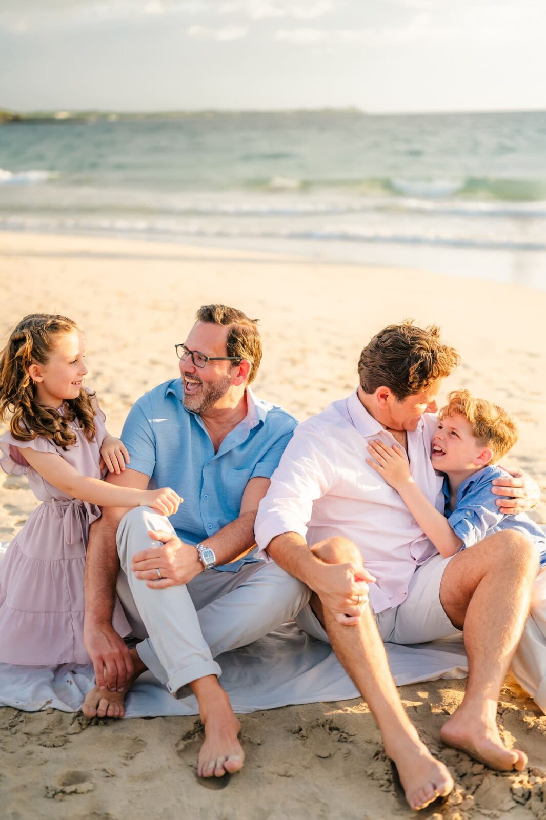 Two men and two children sit together on a blanket at the beach during a Hawaii photo session with kids, smiling and laughing. The ocean waves are in the background under a blue sky. The family appears happy and relaxed.