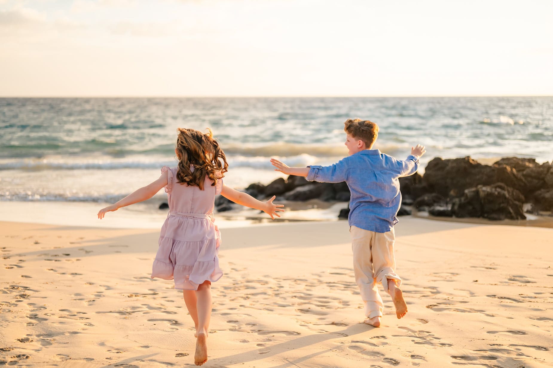 Two children, a girl in a pink dress and a boy in a blue shirt, run barefoot on a sandy beach toward the ocean during a Hawaii photo session with kids, with waves and rocks glowing under golden sunlight in the background.