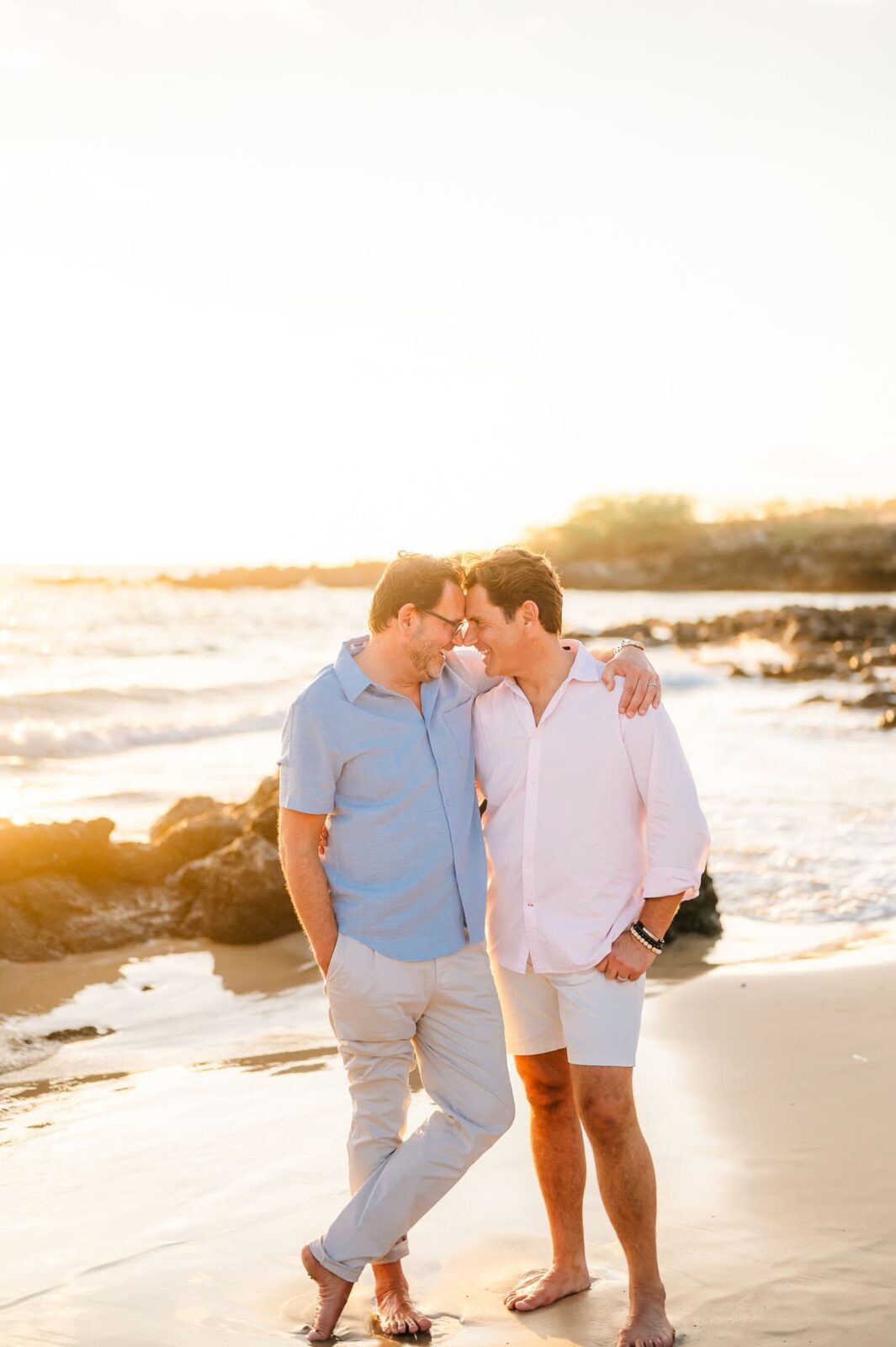 Two men walk barefoot on a sandy beach at sunset, arms around each other and smiling warmly. Waves and rocks glimmer under the golden sky—perfect for a Hawaii photo session with kids capturing family moments by the sea.
