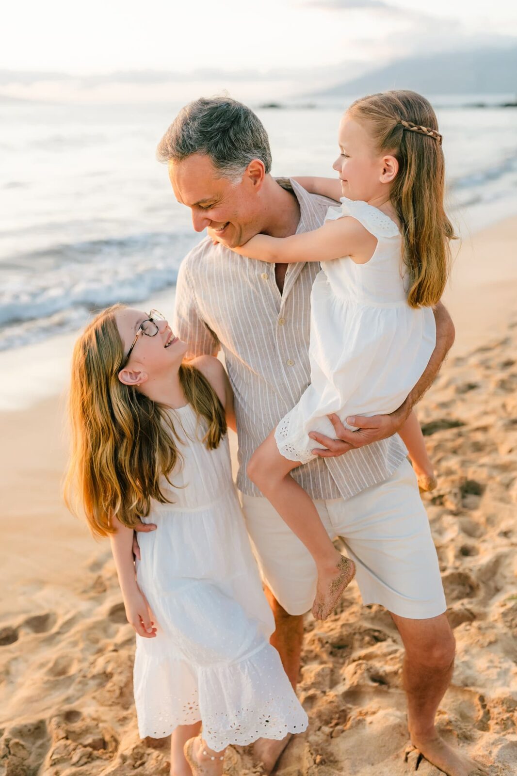 A smiling man stands on a sandy beach at sunset, holding a young girl in a white dress while another girl in a white dress and glasses stands beside him. All three are laughing and looking at each other.