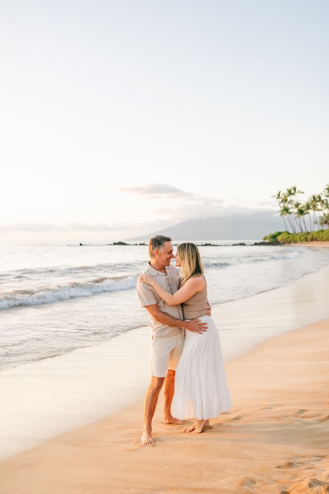 A couple embraces and smiles on a sandy beach at sunset, with gentle waves and palm trees in the background, under a clear sky.