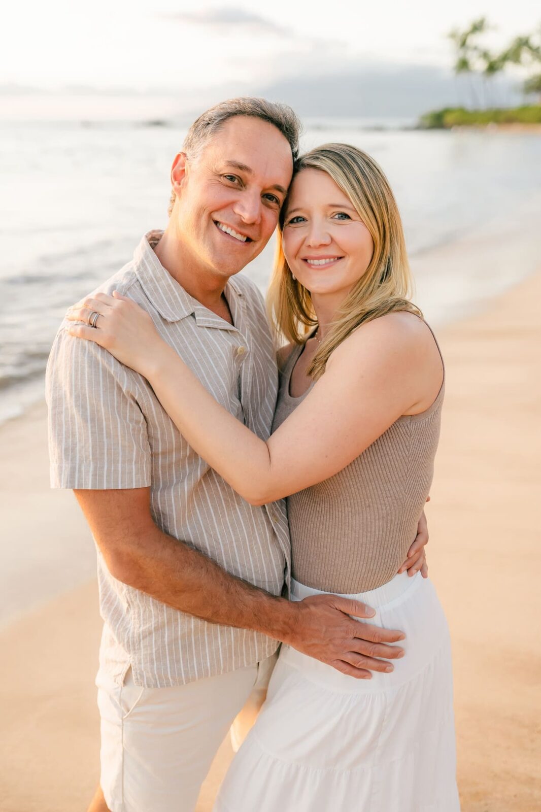 A smiling couple embraces on a sandy beach at sunset, standing close together with the ocean and palm trees in the background. They look happy and relaxed, enjoying the warm, golden light.