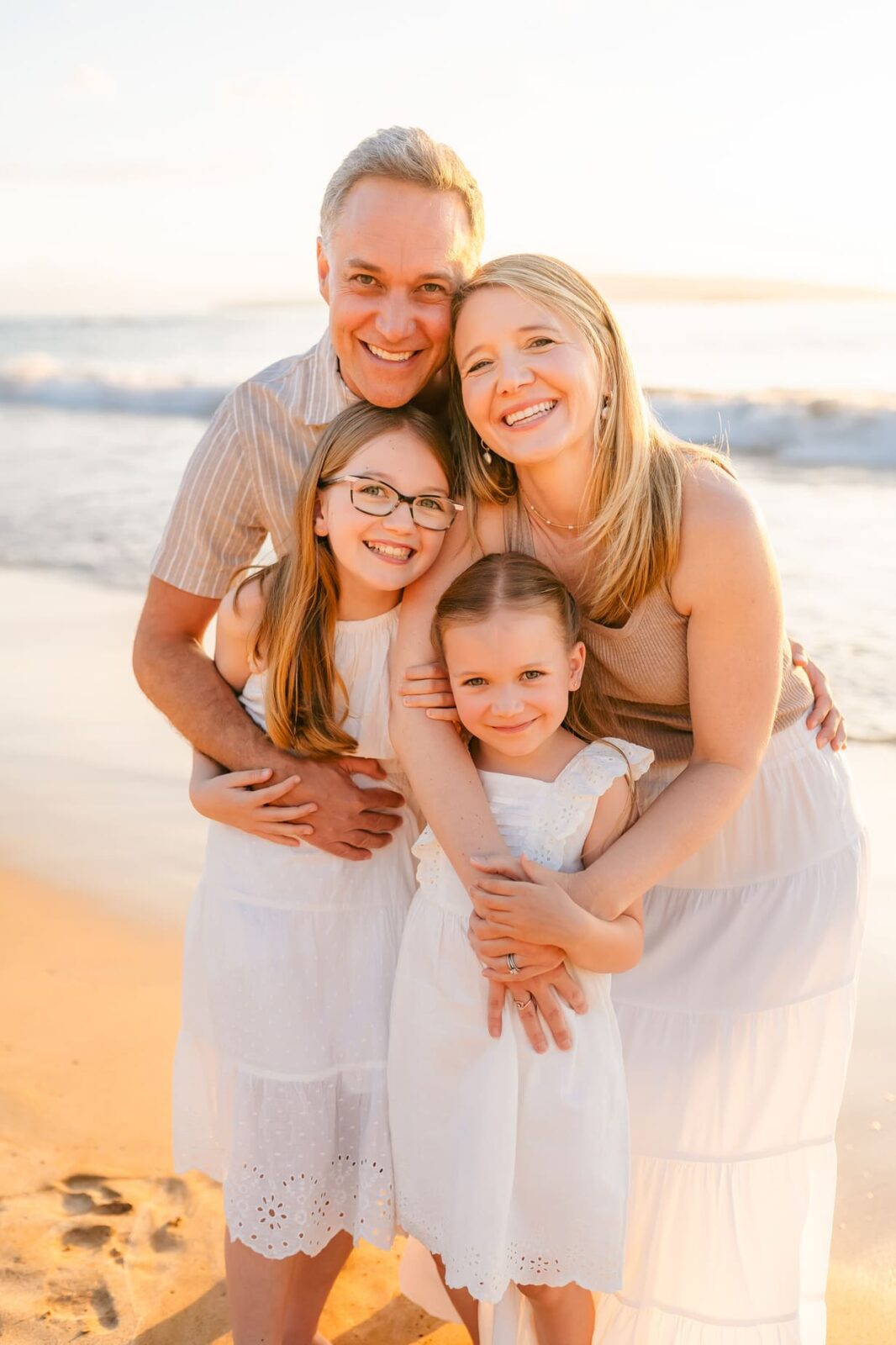 A smiling family of four—two adults and two young girls—stands close together on a sandy beach at sunset, with gentle waves and soft sunlight in the background. All are dressed in light-colored, summery clothes.