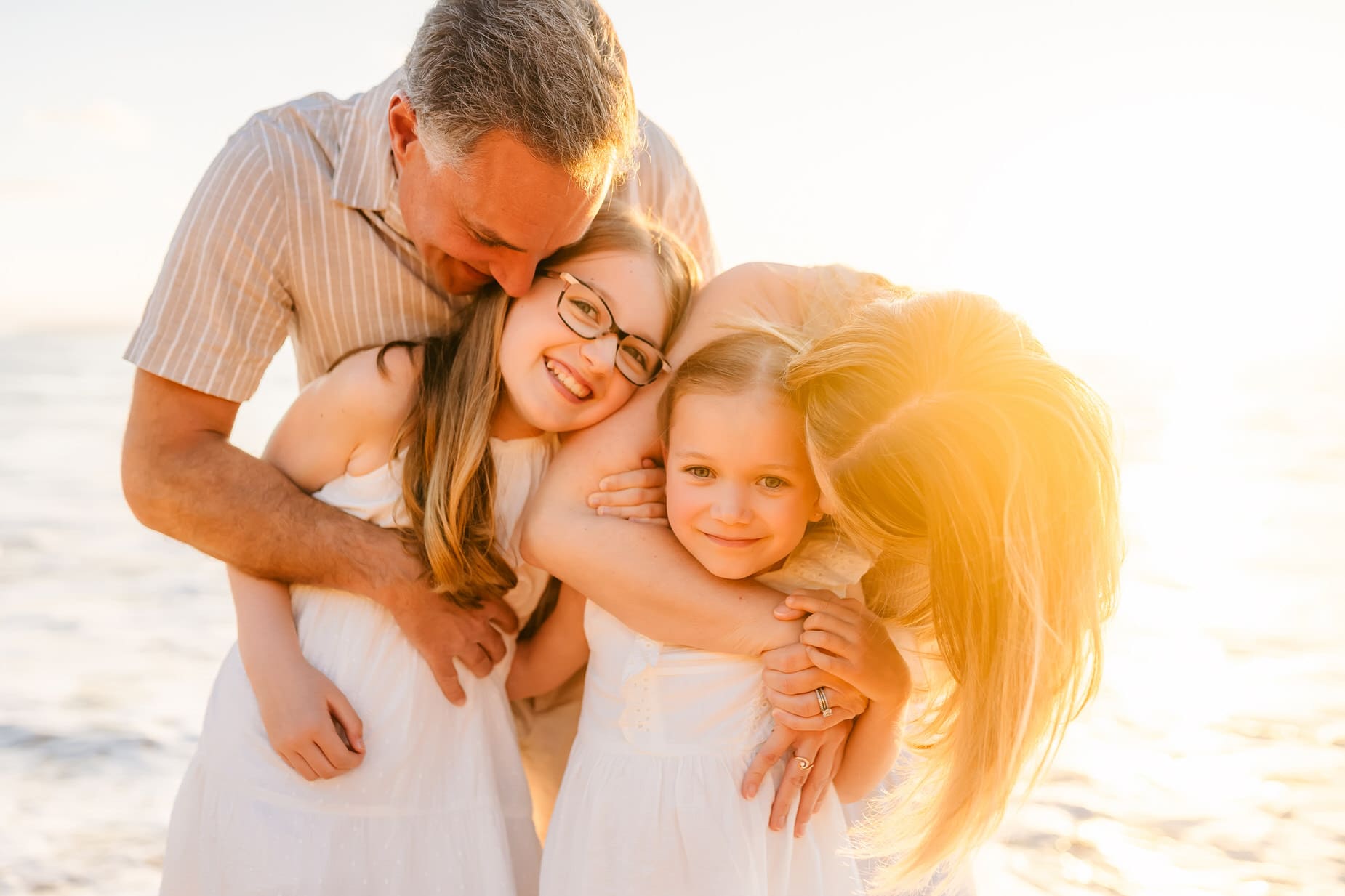A family of four smiles and hugs together on a sunny beach. The parents embrace their two young daughters, who wear white dresses, with the sun shining warmly in the background.