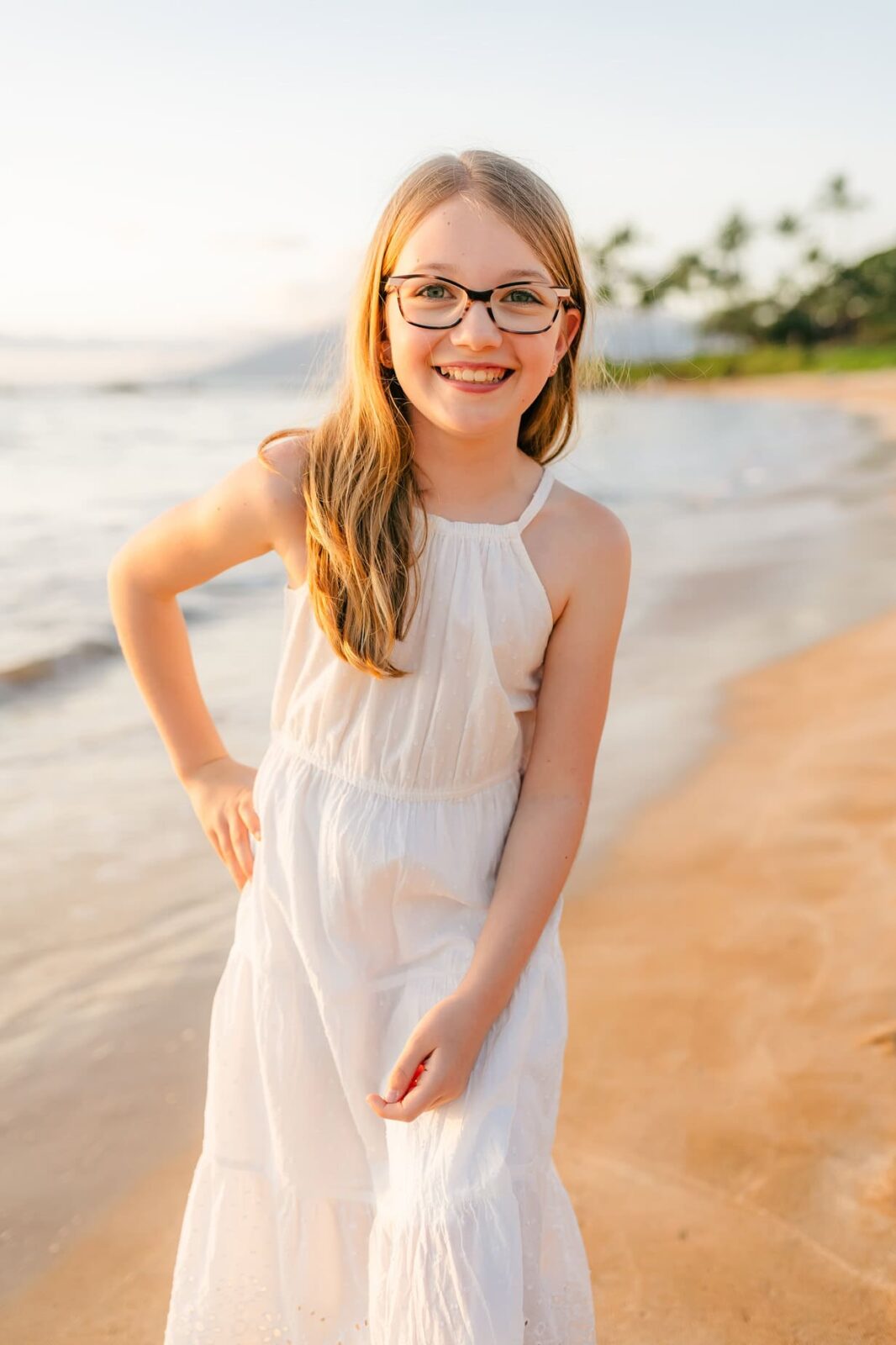 A young girl with long blonde hair and glasses smiles while standing on a sandy beach at sunset, wearing a sleeveless white dress. The ocean and palm trees are visible in the background.