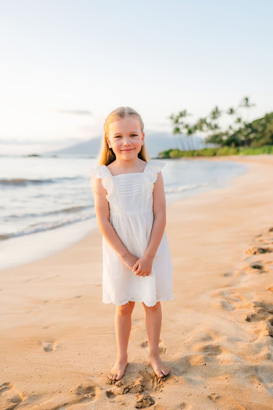 A young girl with blonde hair in pigtails, wearing a white dress, stands barefoot on a sandy beach near the shoreline during sunset, with palm trees and the ocean in the background.
