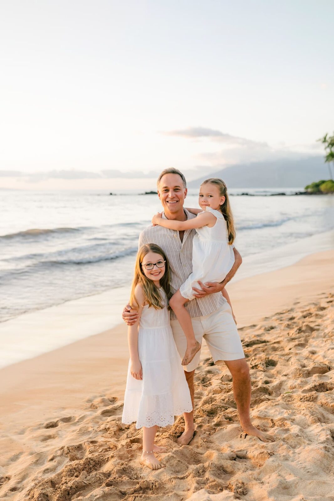 A man stands on a sandy beach at sunset, smiling and holding a young girl in a white dress, while another girl in a matching dress stands beside him. The ocean and sky are calm in the background.