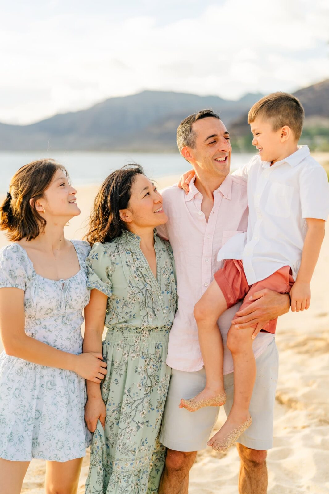 A family of four stands on a sandy beach, smiling and looking at each other. The father holds a young boy, while the mother and daughter stand beside them. Mountains and water are visible in the background.