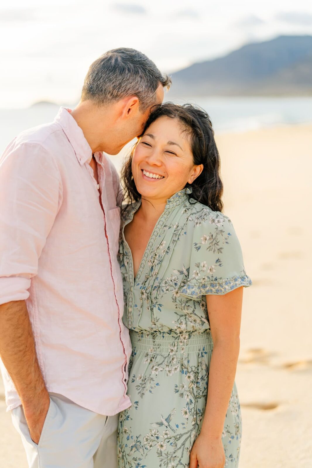A man and woman stand close together on a sandy beach. The man, in a light pink shirt, kisses the woman’s temple. The woman, in a floral dress, smiles warmly. The ocean and distant mountains are visible in the background.