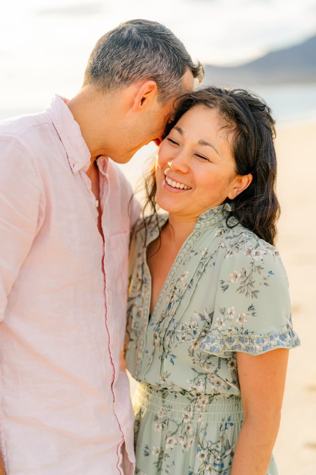 A couple stands close together on a beach at sunset. The man wears a light pink shirt and smiles, leaning his forehead toward the woman, who is smiling and wearing a light green floral dress.