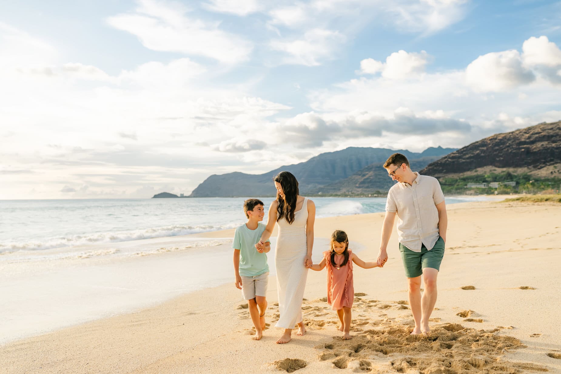 A family of four walks barefoot along a sandy beach, holding hands. The parents and two children enjoy the sunny weather with mountains and ocean waves in the background.