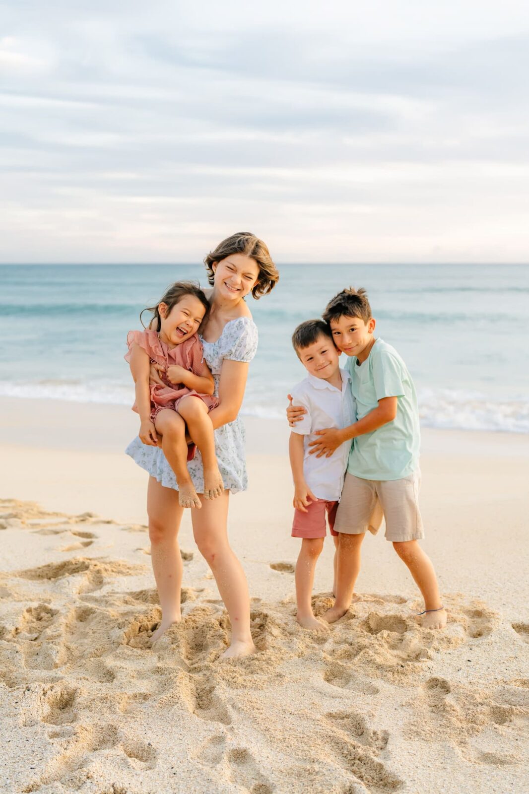A woman stands on a sandy beach holding a young girl, both smiling. Two boys hug each other beside her. The ocean and a cloudy sky are in the background. All appear happy and relaxed.