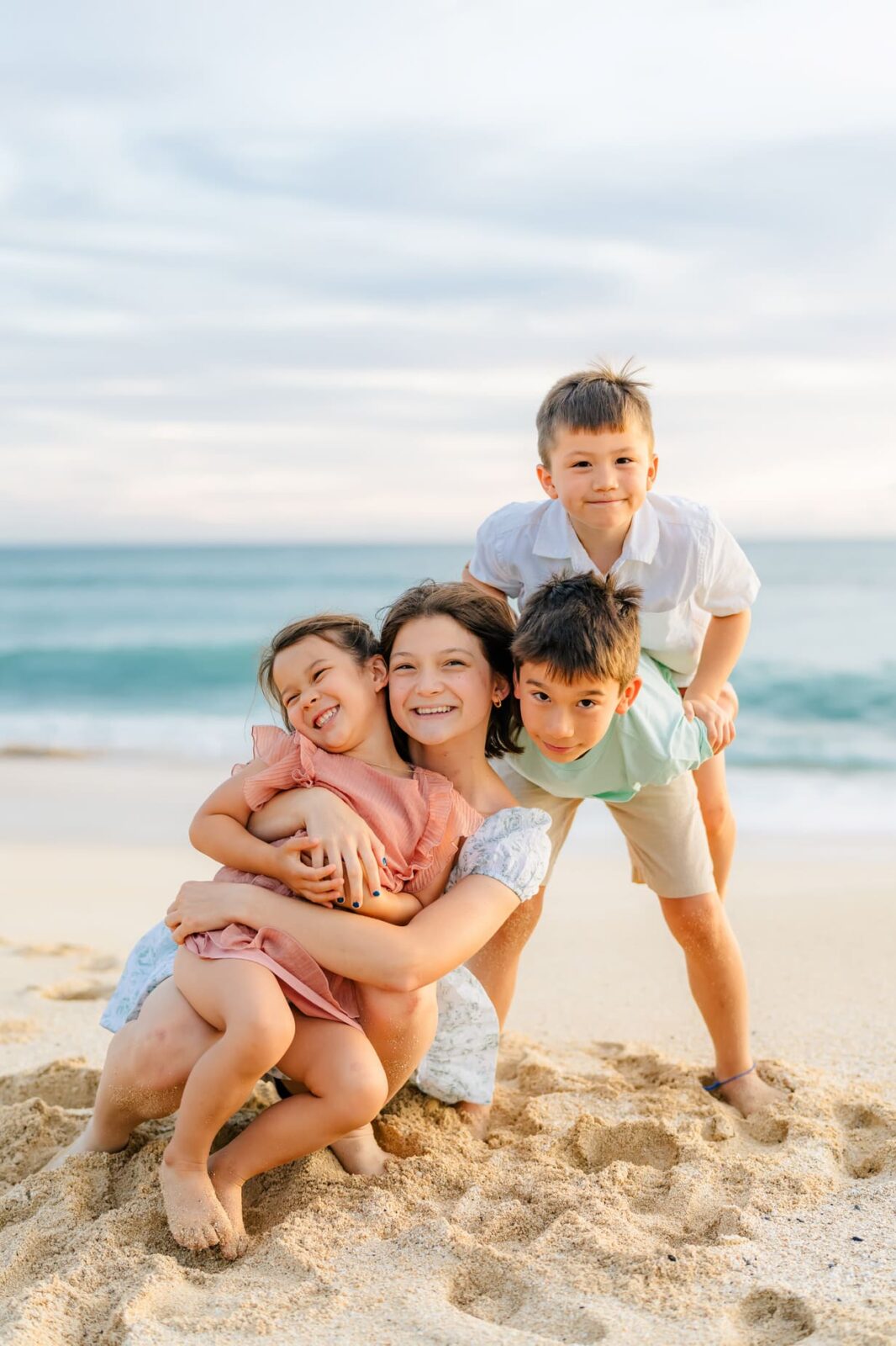 A woman kneels on a sandy beach, smiling and hugging a young girl in a pink dress, while two boys in light-colored clothes pose playfully beside and behind her. The ocean and sky are in the background.