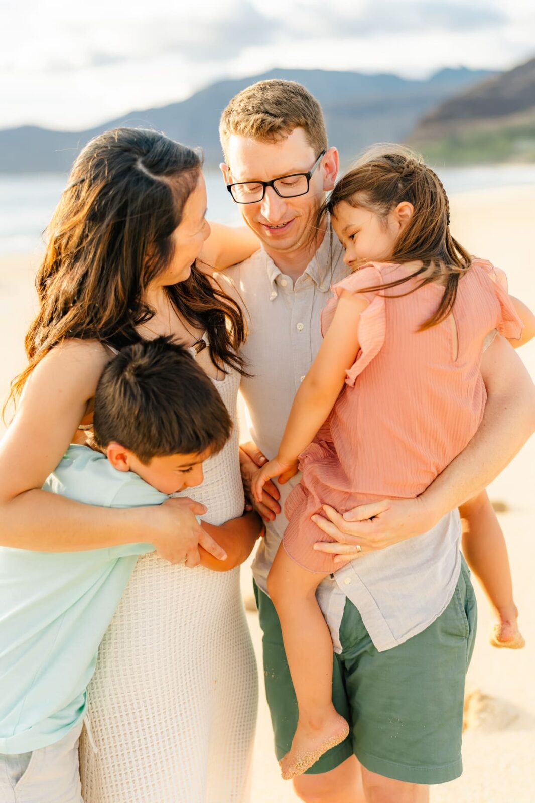 A family of four stands close together on a sunny beach. The parents smile at each other while their young son hugs his mom and their daughter, wearing a peach dress, is held by her dad. Mountains and ocean are in the background.