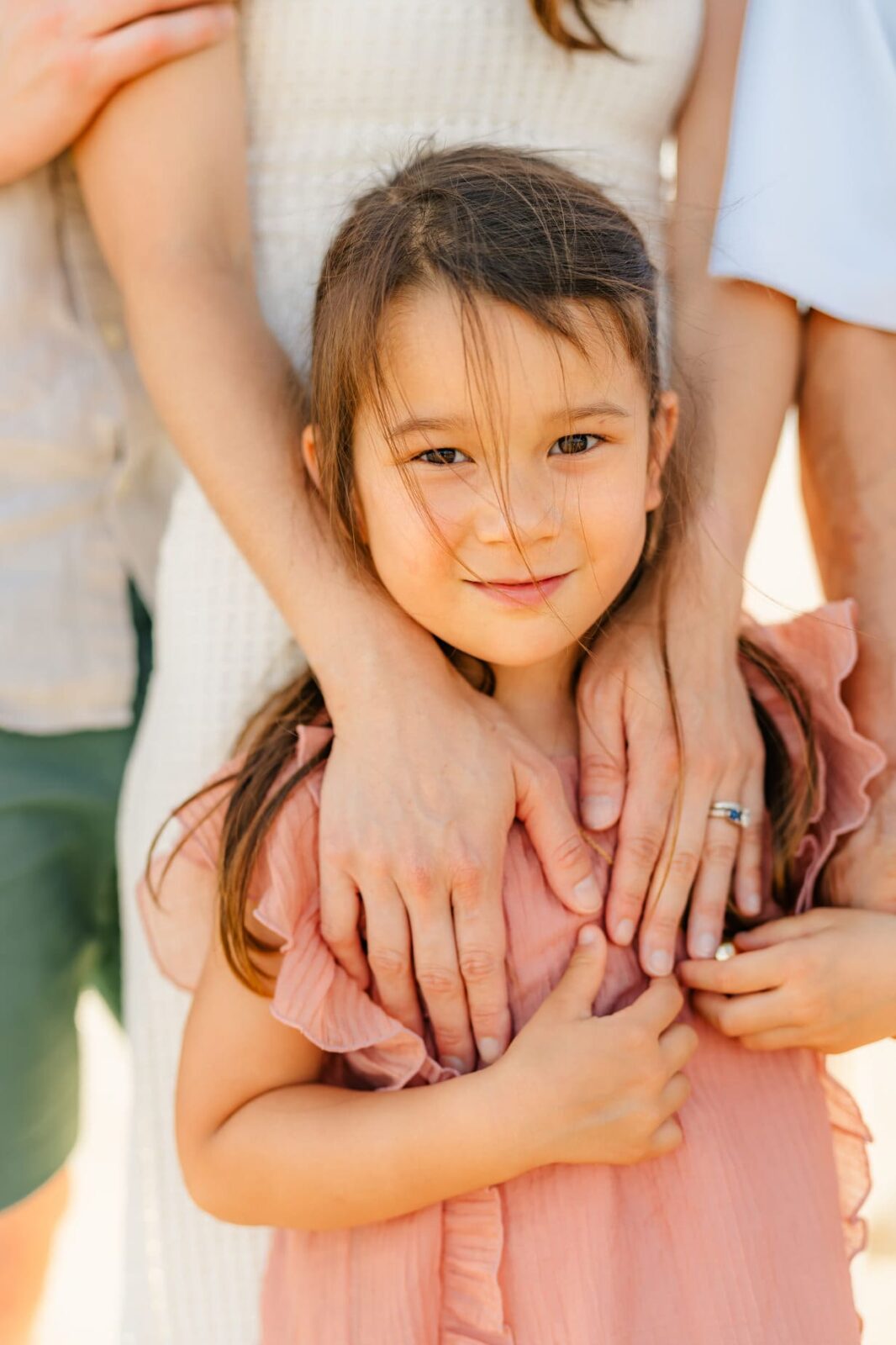 A young girl in a pink dress stands in front of adults, with their hands gently resting on her shoulders. The girl smiles softly at the camera, strands of hair falling over her face. The scene looks warm and affectionate.